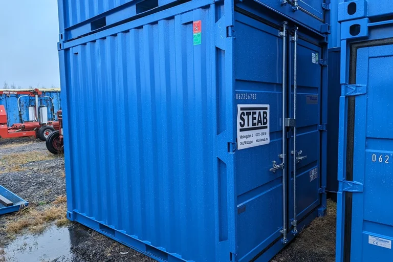 Blå container med STEAB-märkning, stående utomhus på grus och gräs, med utrustning i närheten - inkluderar återköpsgaranti för extra trygghet.