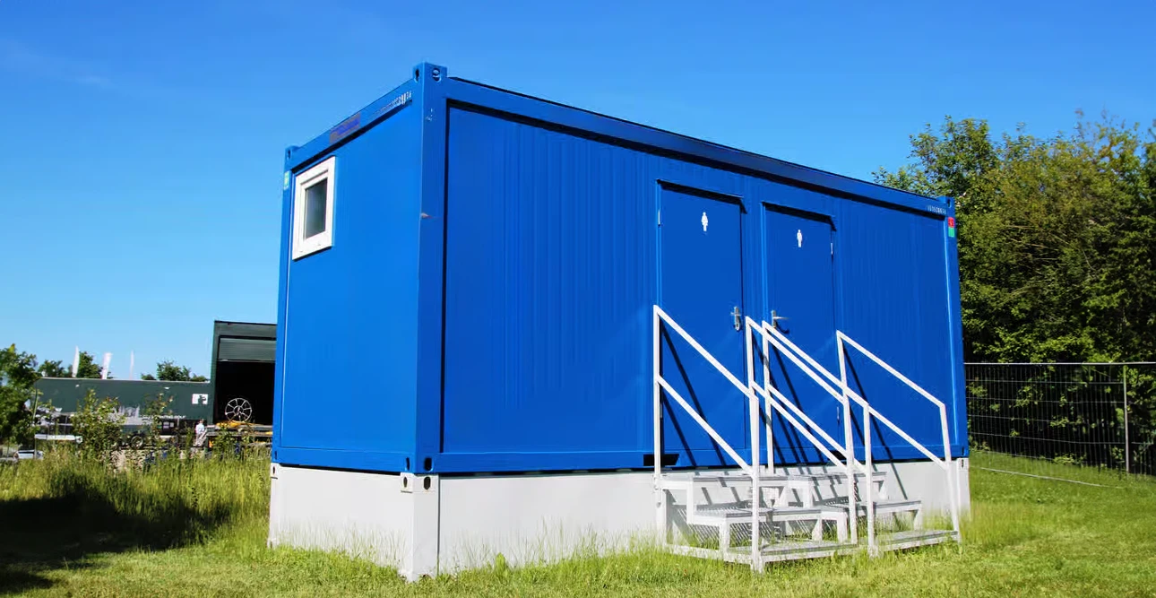 En blå portabel sanitetsmoduler toalettkabin med vita trappor, placerad utomhus på gräs med träd i bakgrunden, perfekt för evenemang som behöver avfallstankar lösningar.