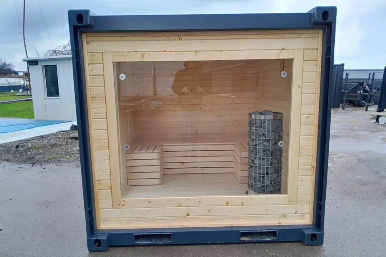 Ett basturum i trä med bänkar och ett bastuaggregat, sett genom ett stort glasfönster i en fraktcontainer.