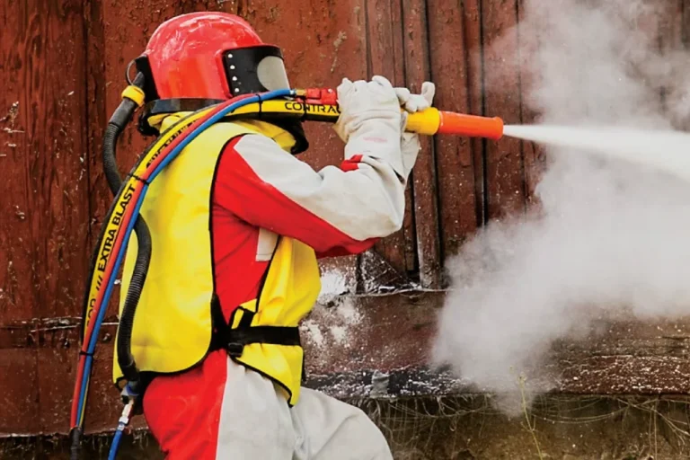 En person i skyddsutrustning använder en högtrycksslang för att spruta på en yta, vilket skapar ett moln av dimma eller damm.
