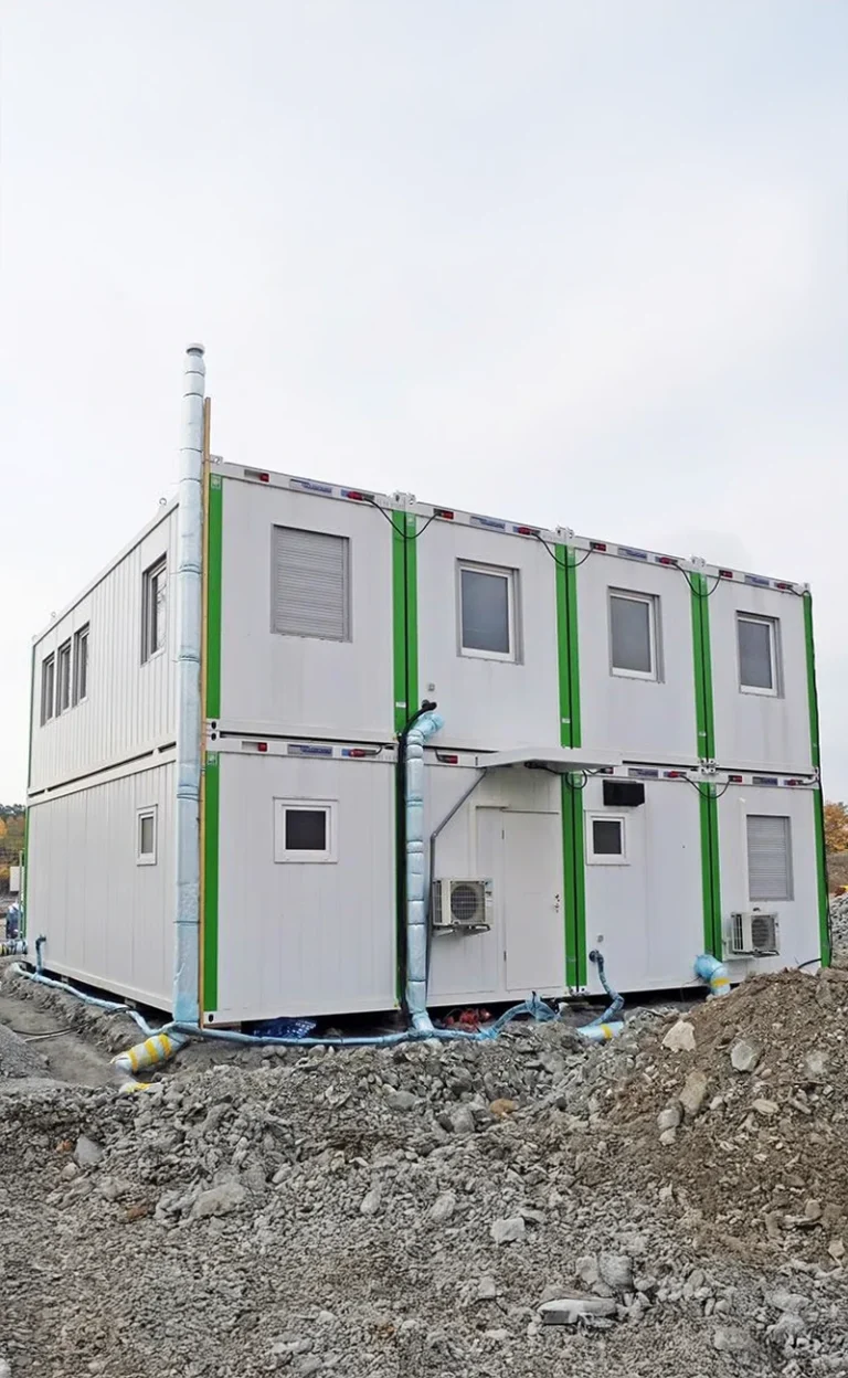 Tvåvånings Byggarbetsplatsmoduler portabel byggnad på en stenig byggarbetsplats, med grön dekor och synliga rörledningar.