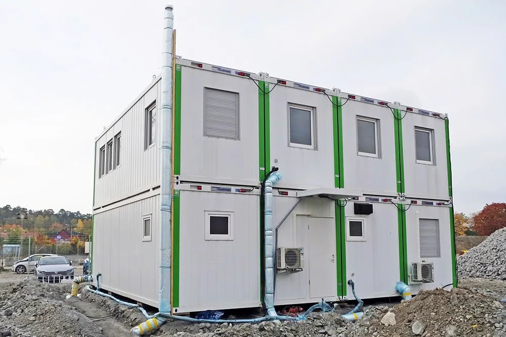 Tvåvåningscontainerbyggnad av Byggarbetsplatsmoduler med gröna detaljer på en byggarbetsplats, bredvid en bil och jordhögar.
