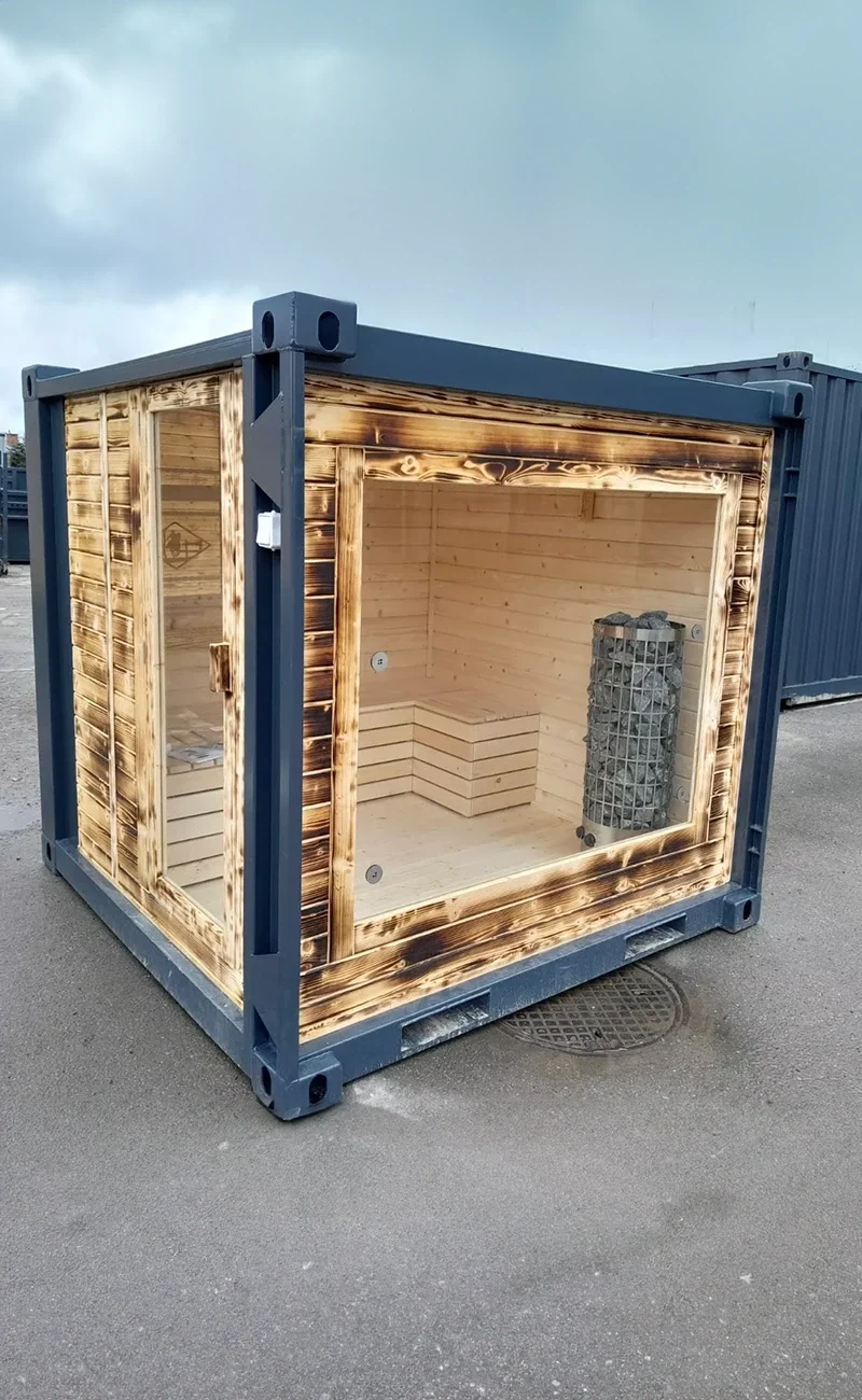 En liten trästuga med stora glasfönster, byggd i en fraktcontainer, står på trottoaren.