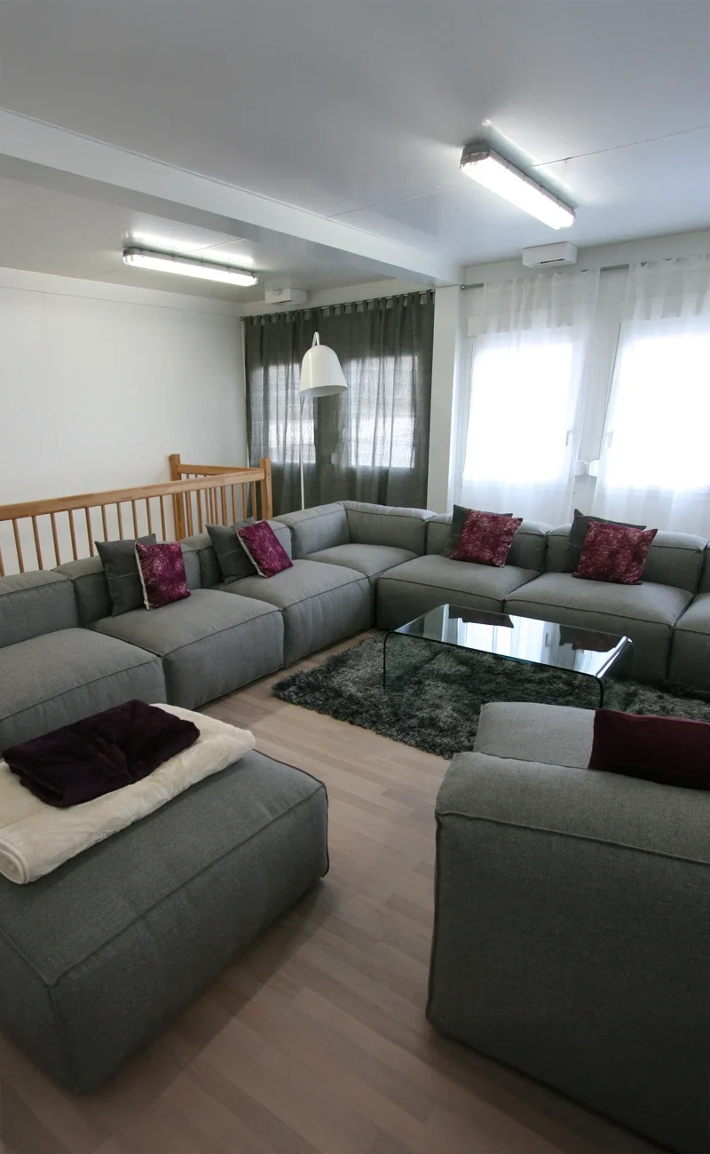 Modernt boende vardagsrum med grå sektionssoffa, lila kuddar, soffbord i glas och stora fönster med gardiner.