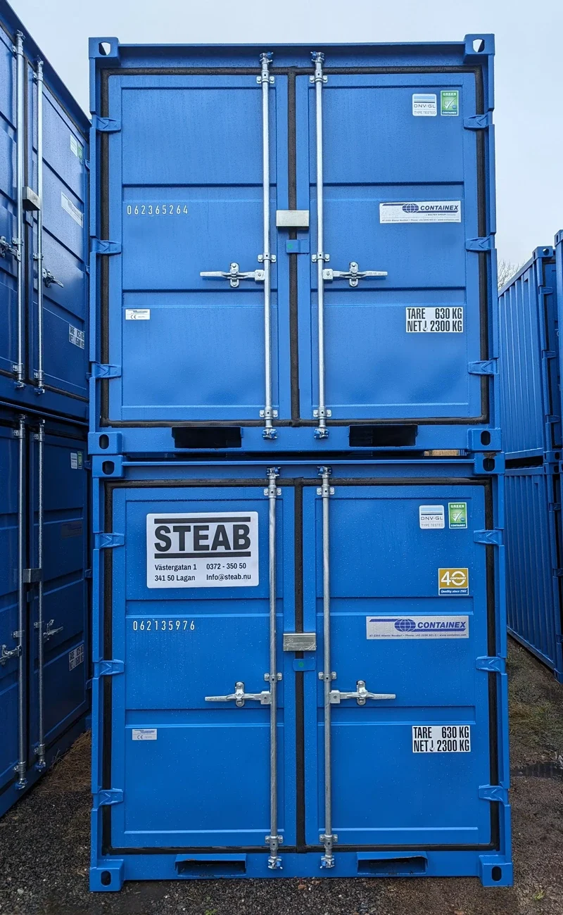 Två staplade blå fraktcontainrar med stängda dörrar och etiketter, omgivna av fler containrar.