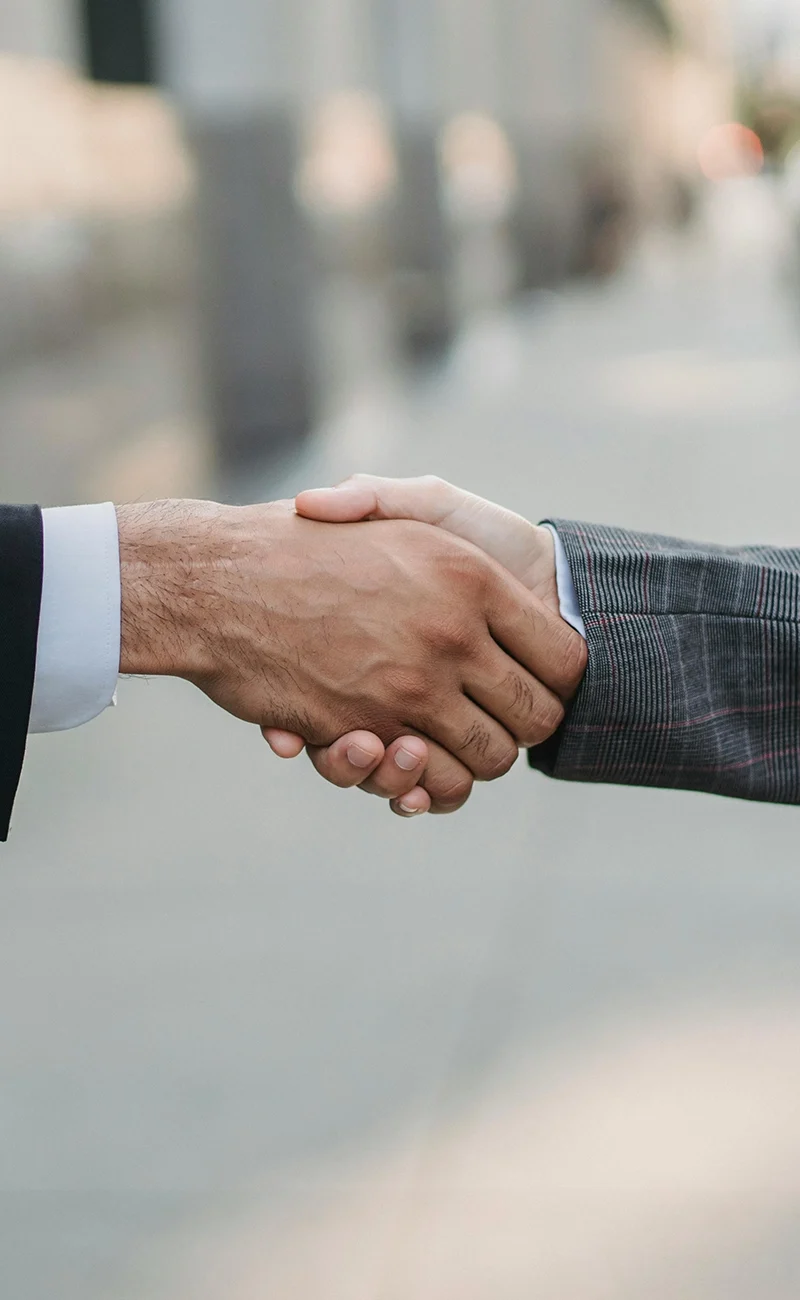 Två kostymklädda personer skakar hand utomhus, symboliserar överenskommelse eller partnerskap, möjligen diskuterar de ulike finansieringsalternativ.