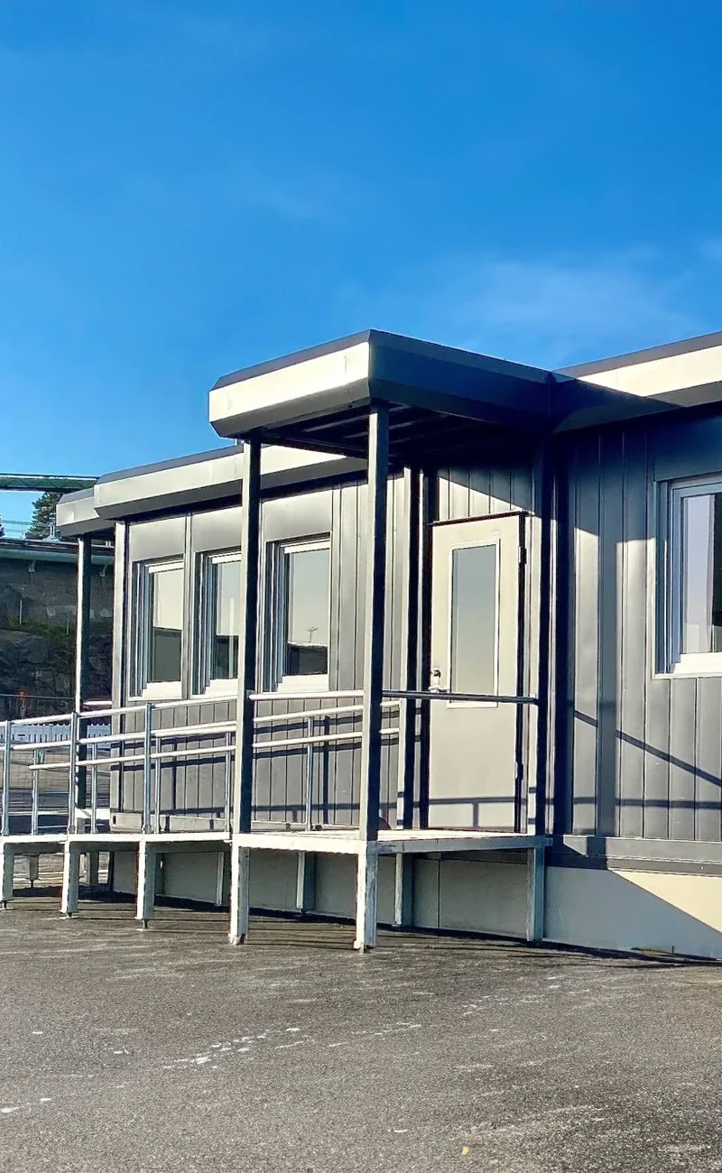 En grå portabel klassrumsbyggnad med fönster, en dörr och en ramp under en klarblå himmel - perfekt för flexibla utrymmen som kontorsmoduler.