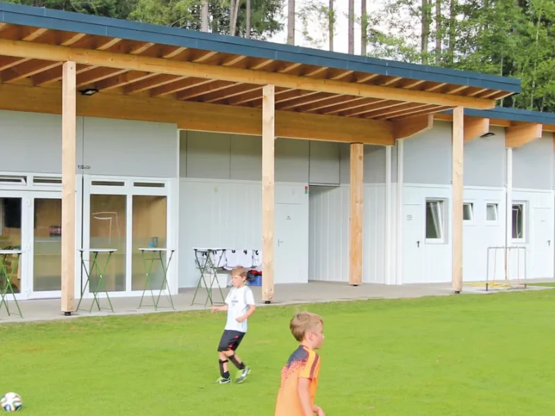Två pojkar spelar fotboll på gräset framför en modern byggnad med stora fönster, träbjälkar och ett närliggande omklädningsrum där spelarna kan byta om.