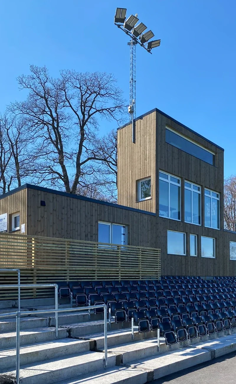 Modern sportpaviljong i trä med stora fönster, stadionbelysning, tomma rader av svarta stolar och bekvämt omklädningsrum under en blå himmel.