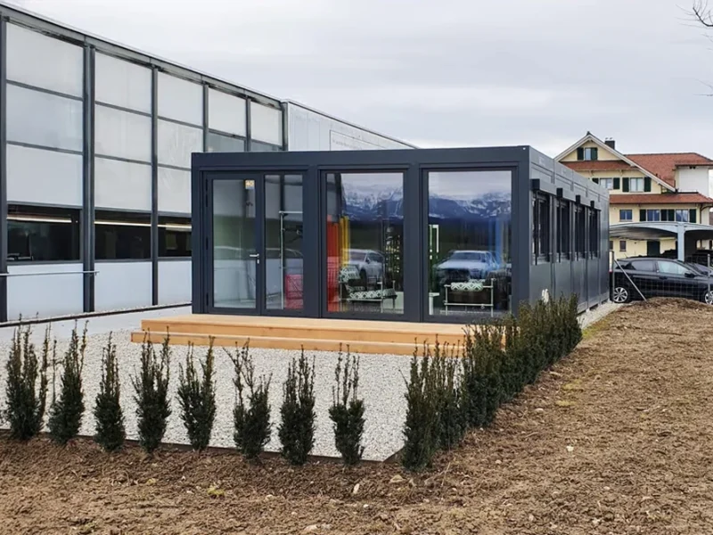 Modern modulbyggnad med stora fönster fungerar som ett elegant showroom, omgivet av grus, små buskar och bilar i närheten - en perfekt miljö för att visa upp glasmoduleringslösningar.