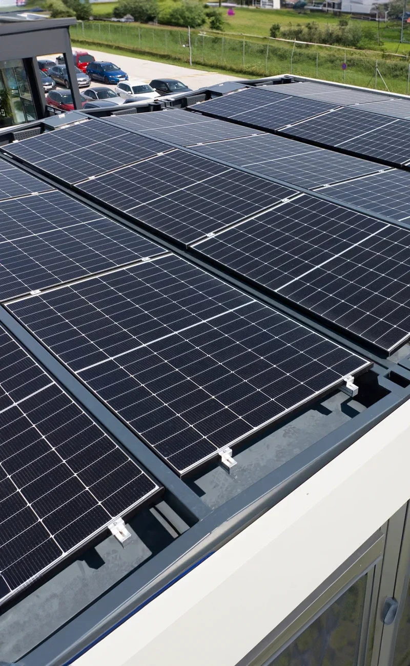 Rader av solcellsmoduler installerade på ett hustak, med parkerade bilar, byggnader och en containrar i bakgrunden.