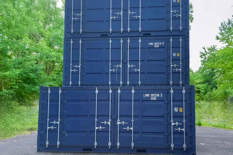 Tre stora blå Miljöcontainer-fraktcontainrar staplade utomhus, omgivna av gröna träd och gräs.