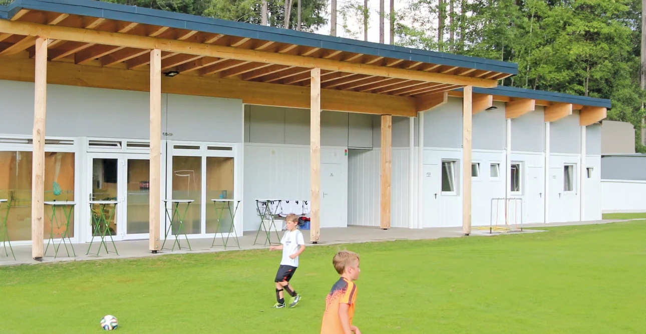 Två pojkar spelar fotboll på gräset framför en modern vit byggnad med träbjälkar, stora fönster och en omklädningsrum i närheten.