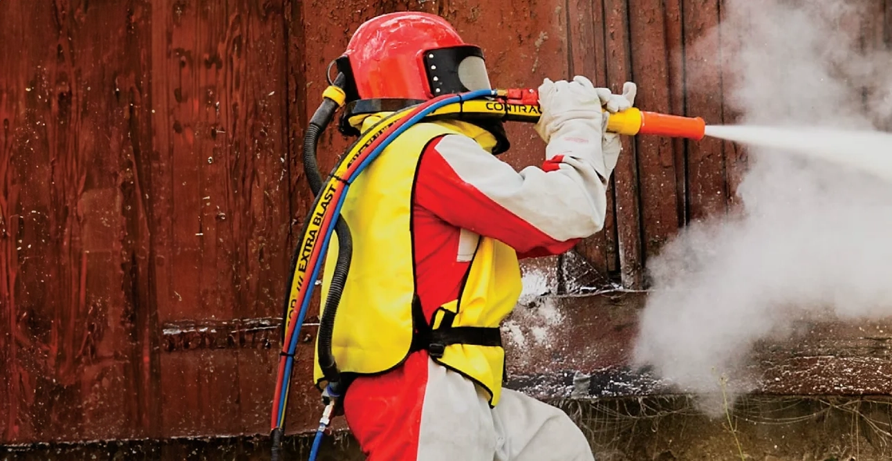 En person i skyddsutrustning använder en slang för att spruta dimma eller vatten mot en rostig metallyta.