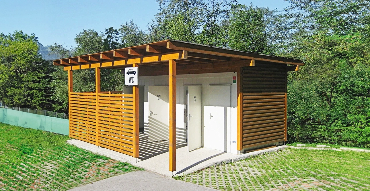 Offentlig toalettanläggning i trä med två vita dörrar och WC-skylt, omgiven av grönska - perfekt för parker som behöver moderna Toalett- eller Duschmoduler-lösningar.